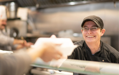 Cafeteria staff member handing over food to a customer with a big smile